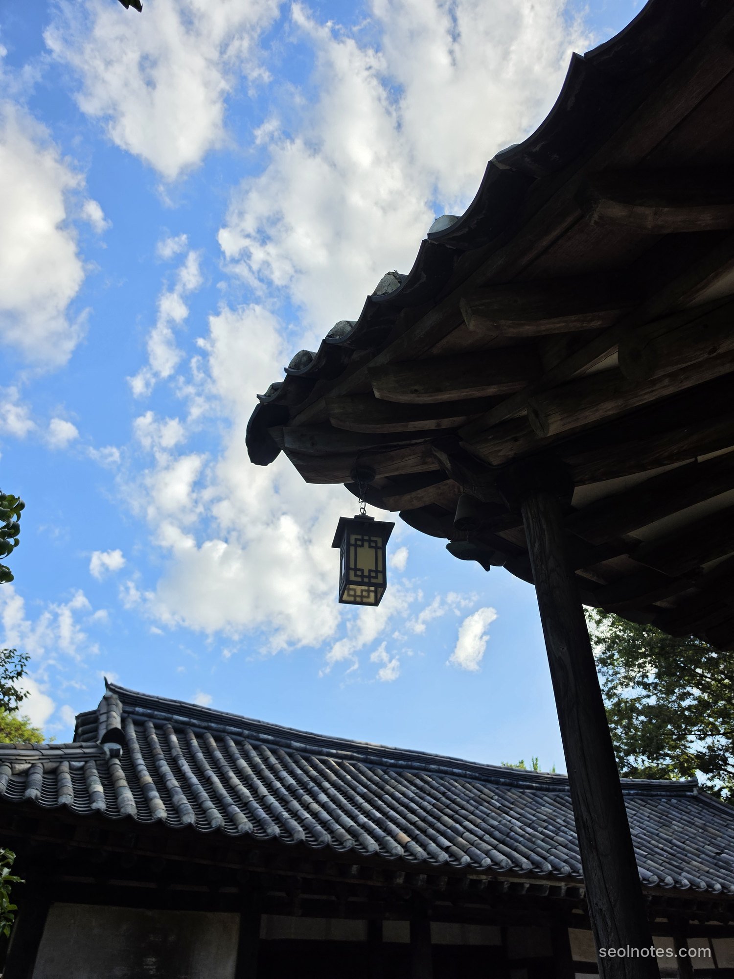 Hanok eaves and a lantern against a cloudy blue sky