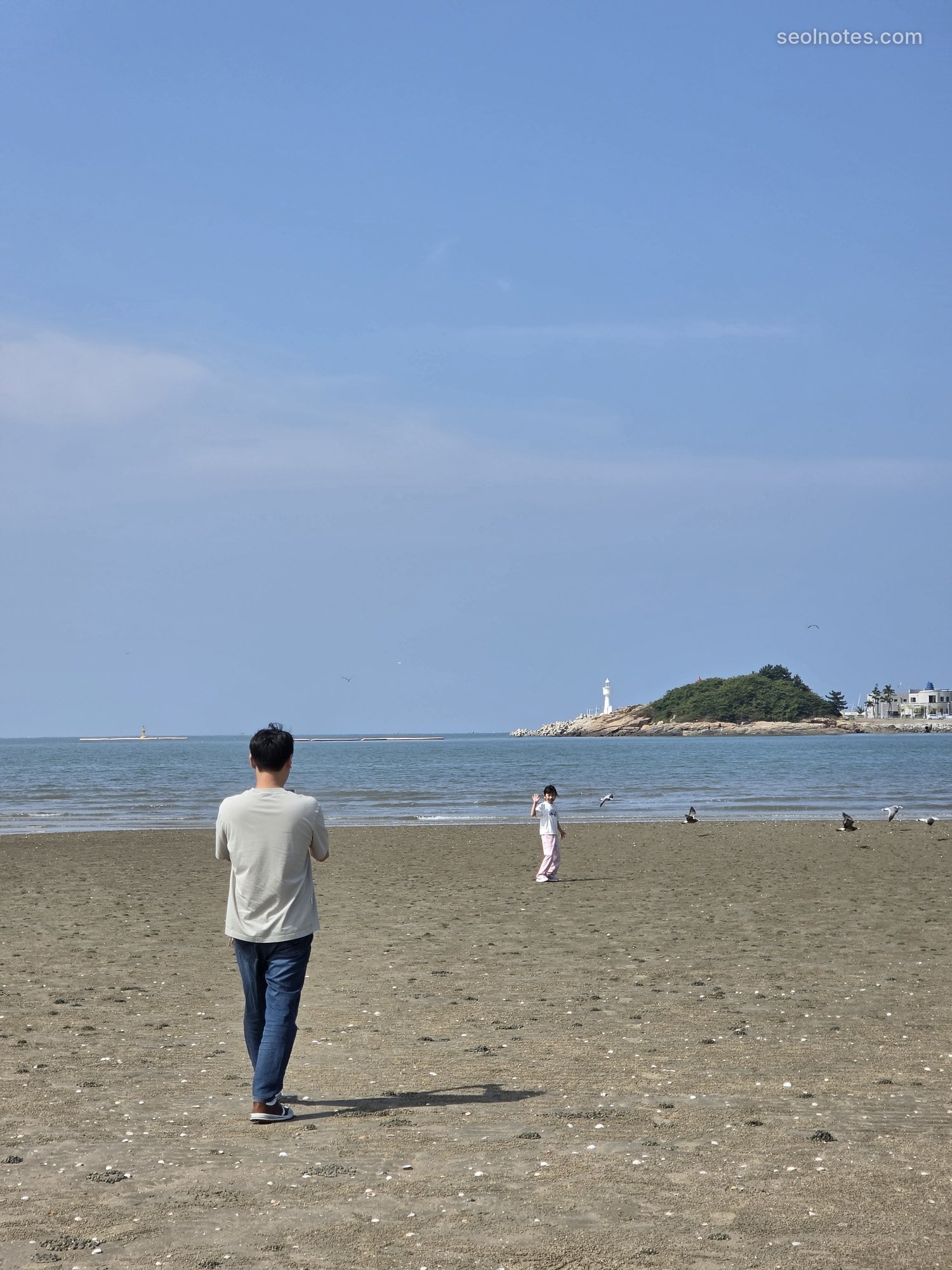 A man walking on a wide sandy beach toward a small lighthouse island