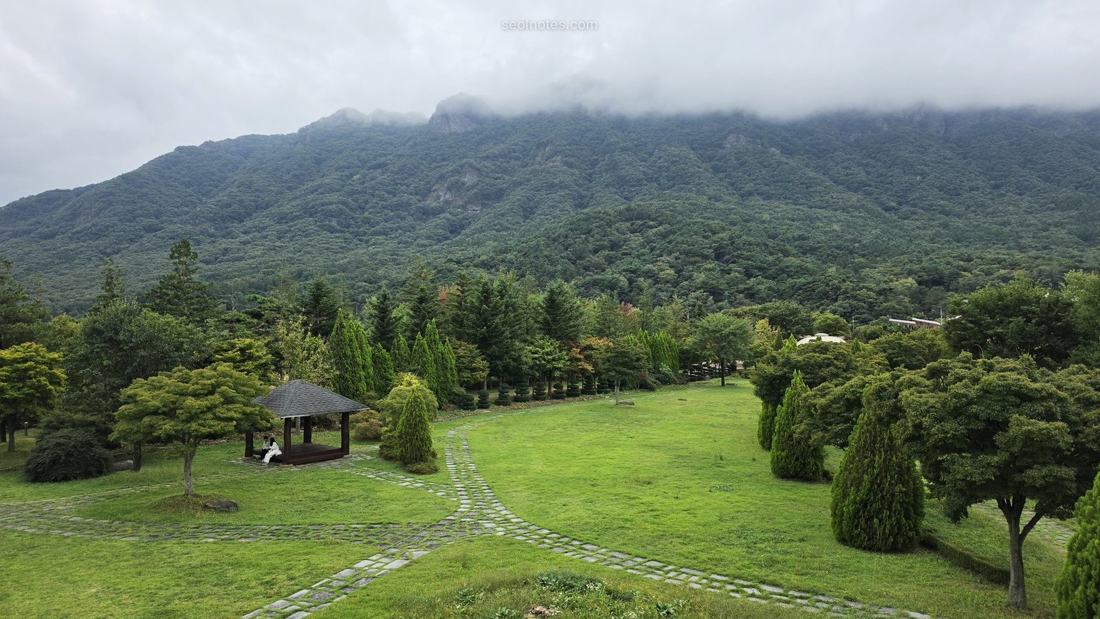 A green lawn and pavilion below a cloud-topped mountain